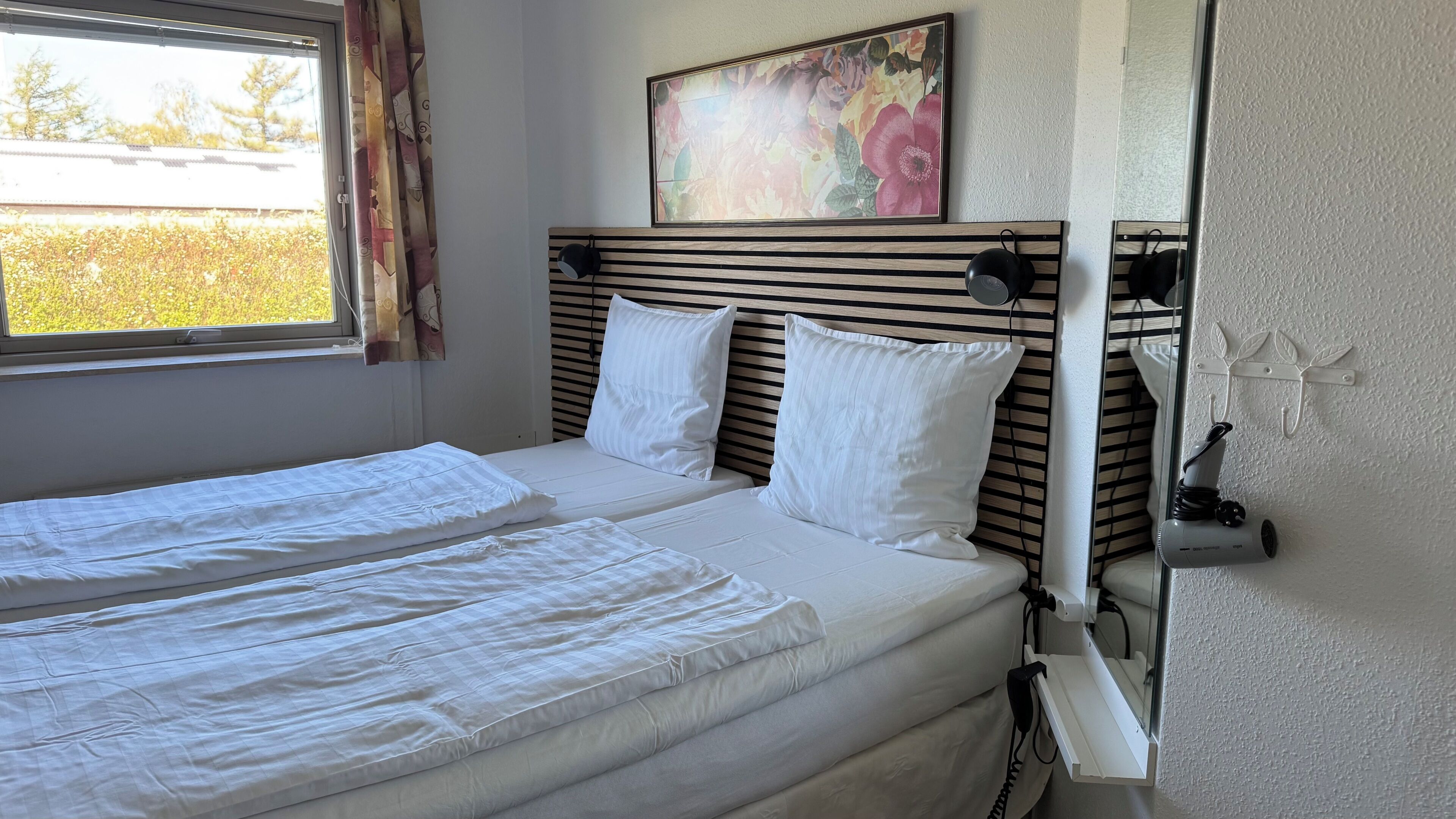 Een tweepersoonsbed met witte lakens, een hoofdbord met horizontale latten, een raam met gordijnen, en een bloemrijk schilderij boven het bed.