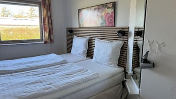 Een tweepersoonsbed met witte lakens, een hoofdbord met horizontale latten, een raam met gordijnen, en een bloemrijk schilderij boven het bed.