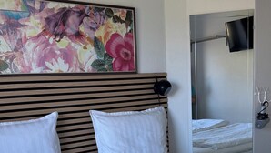 A hotel room with a large bed, a headboard with a floral painting, a bedside table with a lamp, and a television mounted on the wall. - Hine Bakke Guest House (Rudkobing)