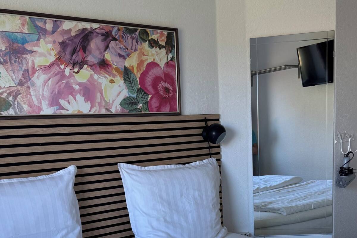 Ein Hotelzimmer mit einem großen Bett, einem mit floralem Gemälde verzierten Kopfteil, einem Nachttisch mit Lampe sowie einem an der Wand befestigten Fernseher.
