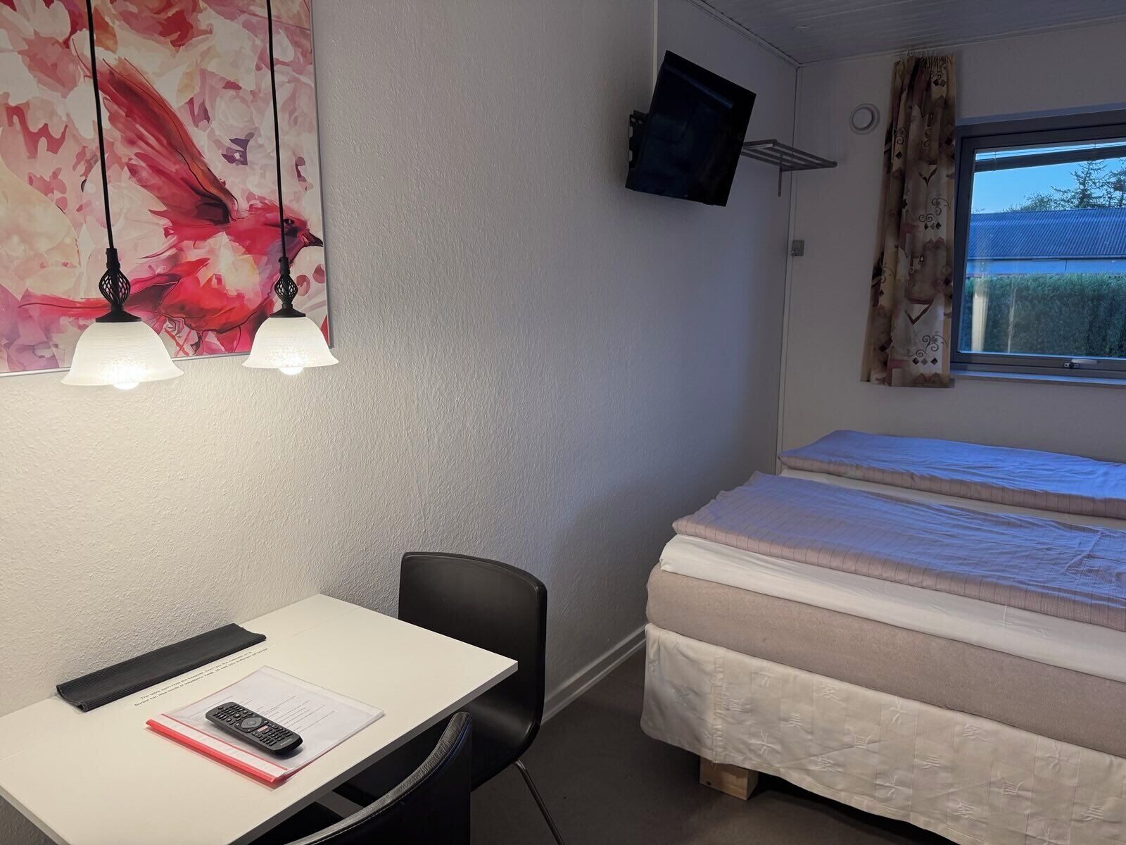 Un dormitorio con cama, escritorio y silla, televisor montado en la pared y ventana con cortinas.