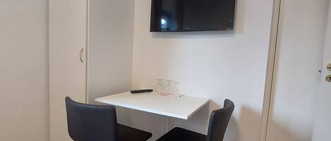 Una habitación pequeña y moderna con un escritorio blanco, dos sillas negras, un televisor de pantalla plana montado en la pared y un armario blanco.