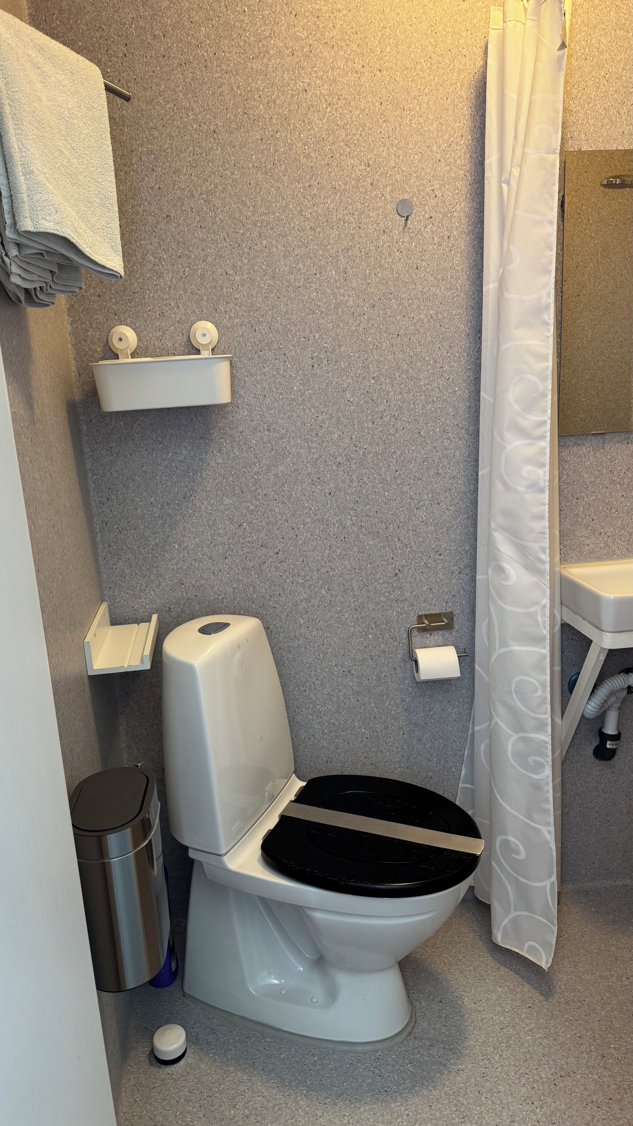 A bathroom with a toilet, a trash can, a towel rack, a shelf, a shower curtain, and a sink.