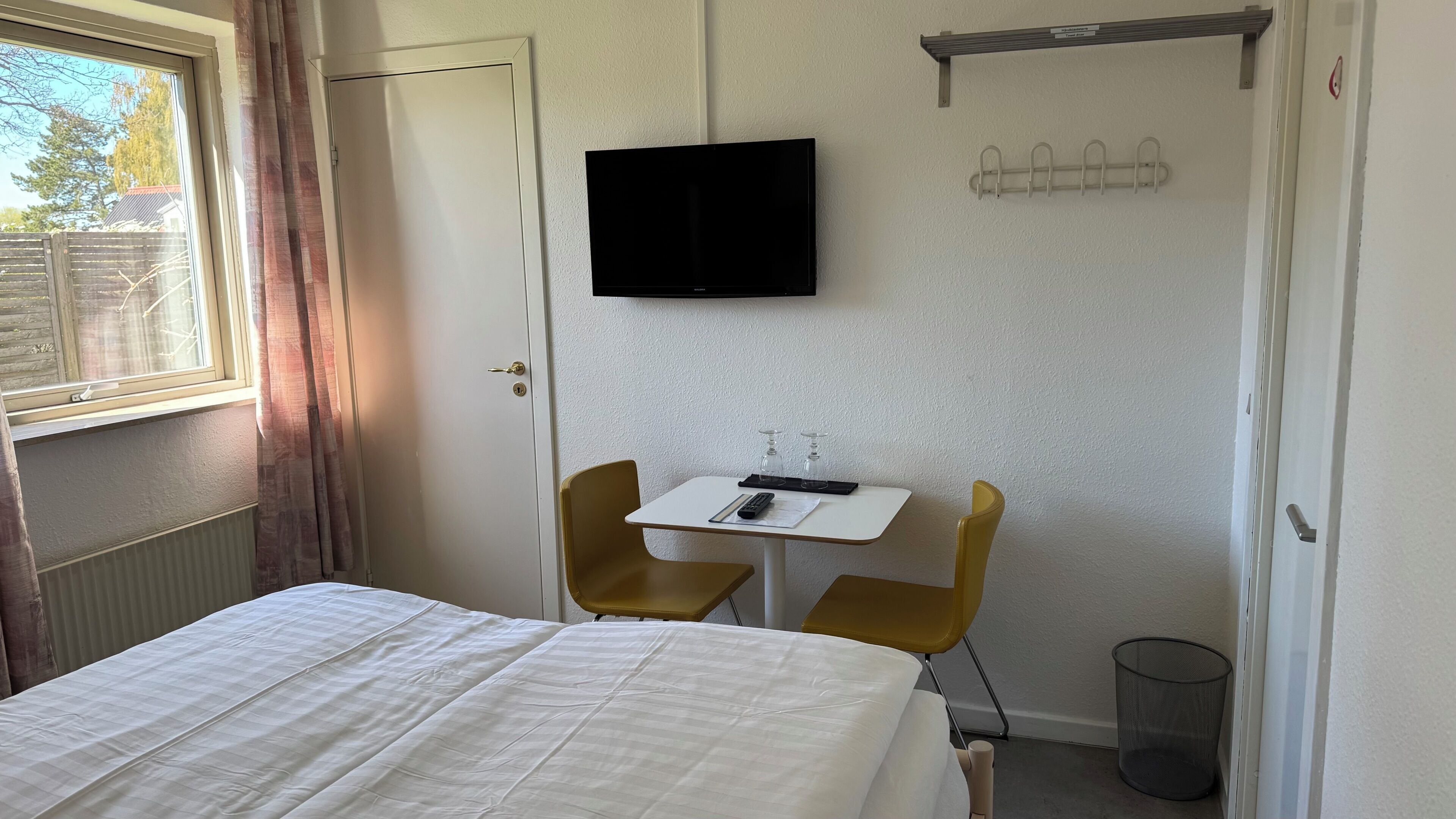 A room with a bed, a small table with a chair, a television mounted on the wall, and a coat rack.