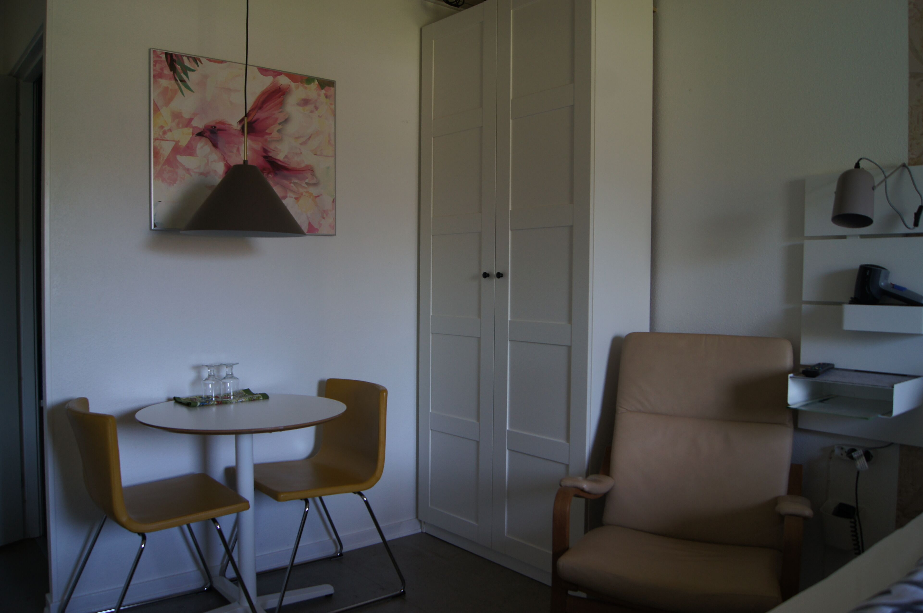 Une pièce avec une armoire blanche, une table ronde accompagnée de deux chaises, un fauteuil beige et un tableau accroché au mur.