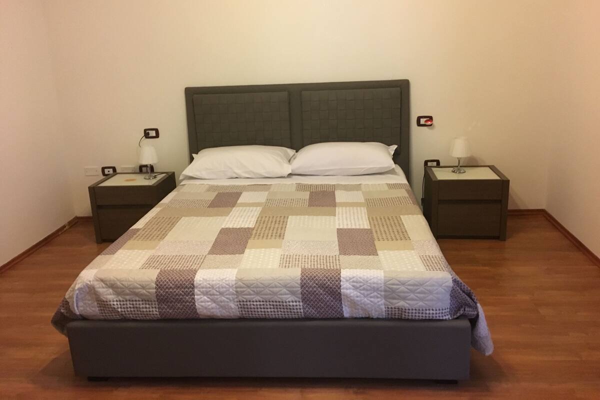 Una cama bien hecha con colcha, dos mesitas de noche con lámparas y un reloj de pared.