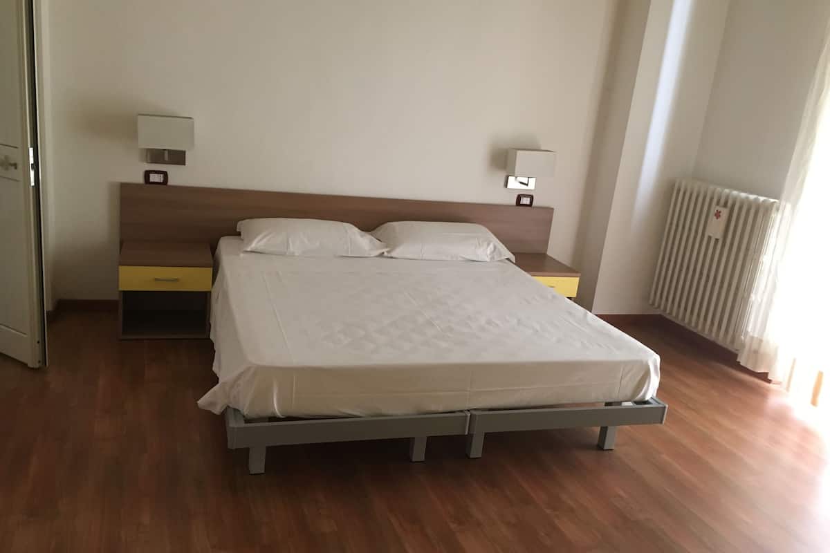 Una cama bien hecha en una habitación de hotel con cabecera de madera, una mesita de noche con lámpara y un radiador.