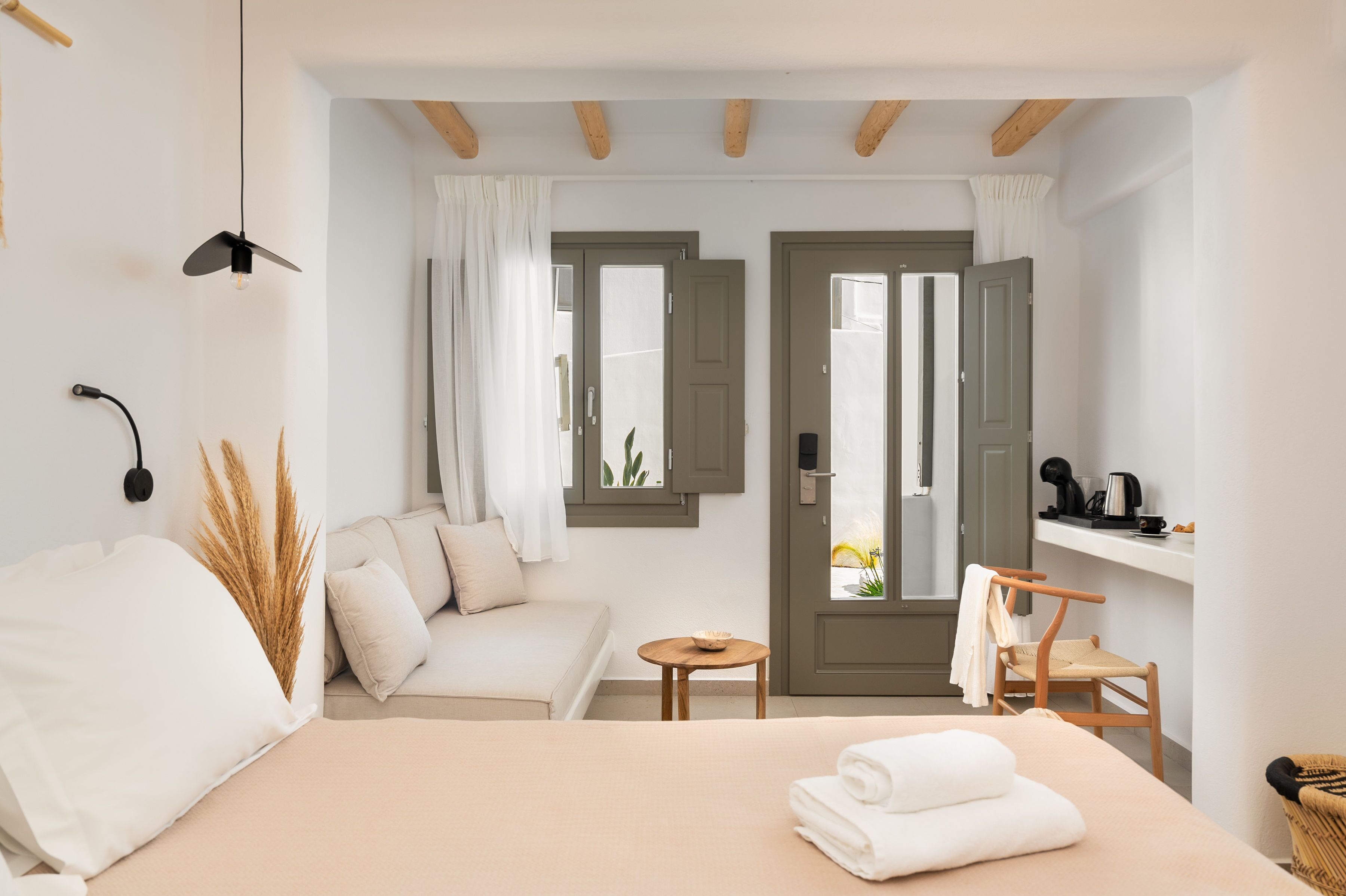 Ein modernes Hotelzimmer mit Bett, Sofa, kleinem Tisch und Stuhl. Zwei Türen führen zu einem Außenbereich mit Blick auf Blumen.