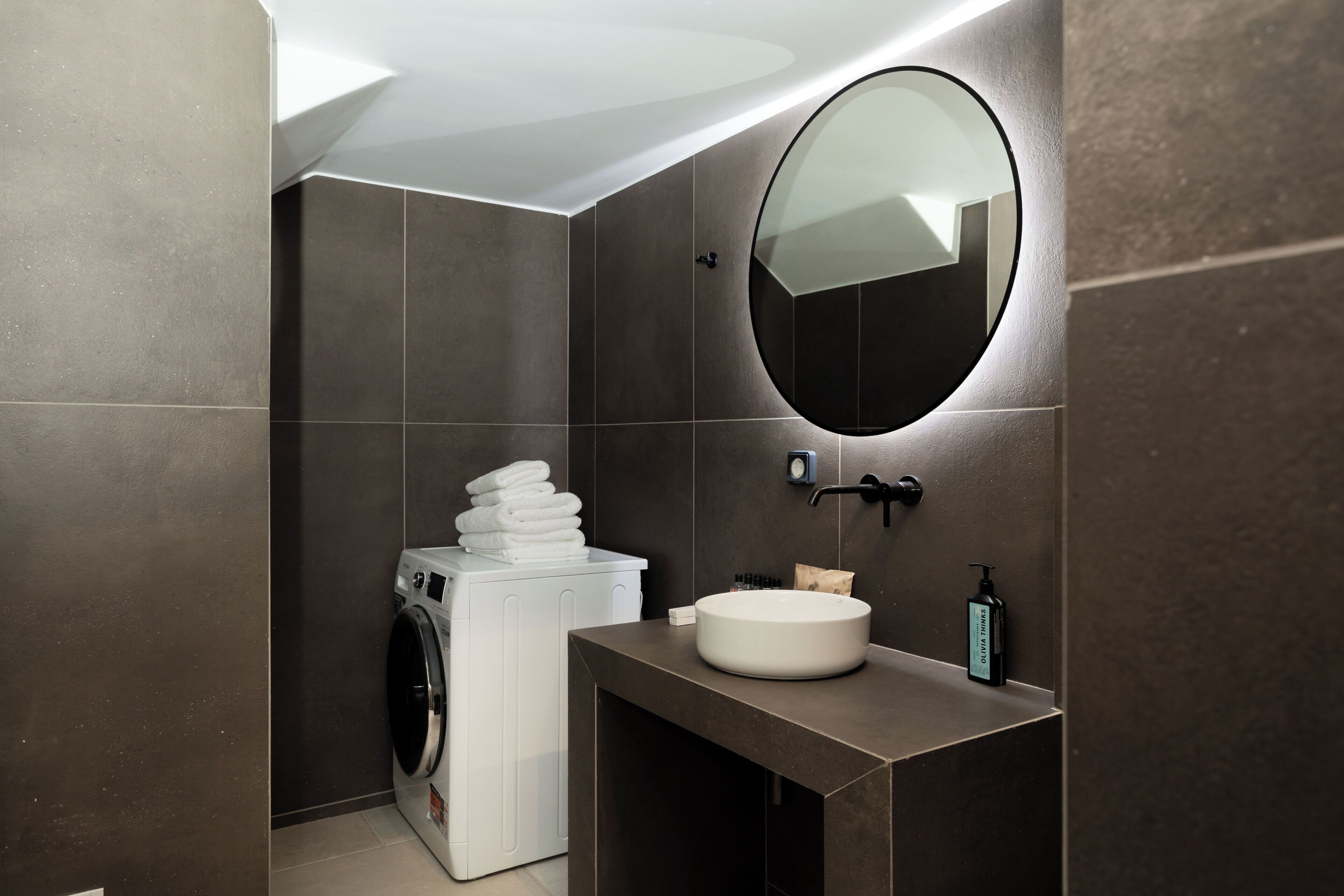 Un baño moderno con lavadora integrada, un espejo redondo y un lavabo con artículos de aseo.