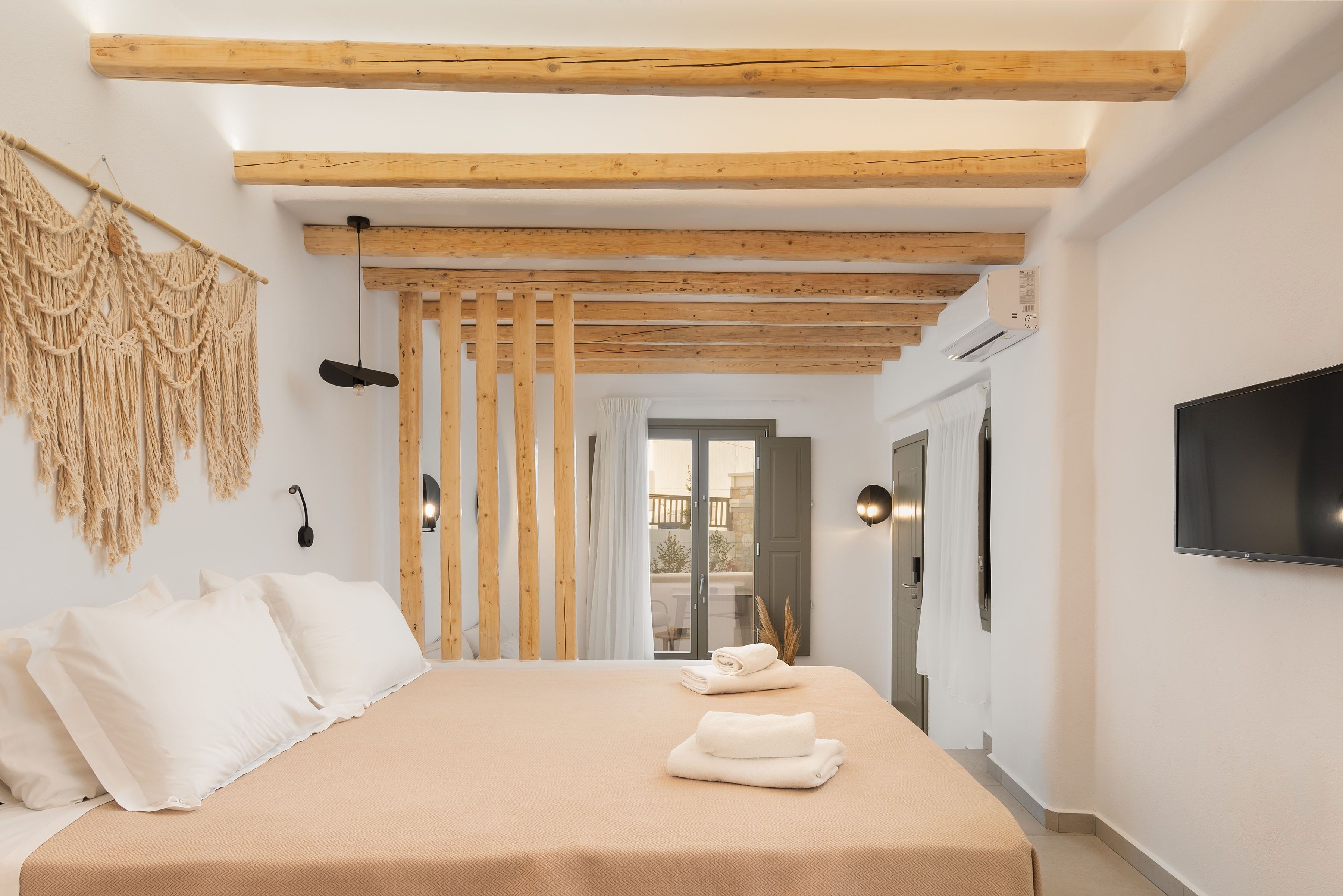 Un dormitorio con techo de madera, cama con ropa de cama blanca, televisor en la pared y puerta al balcón.