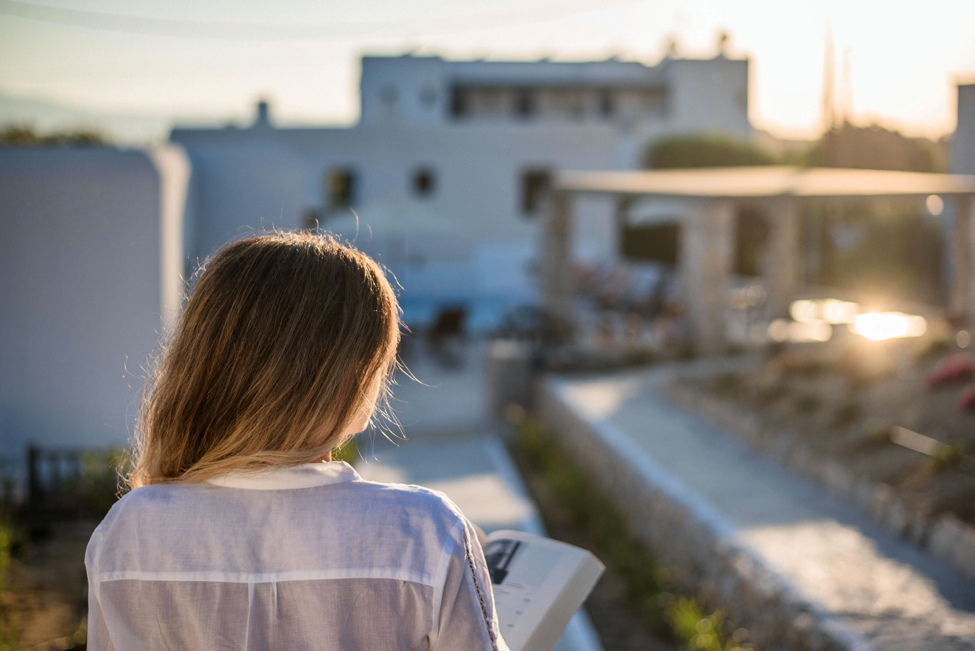 A person reading a book on a balcony overlooking a residential area with buildings and a clear sky.