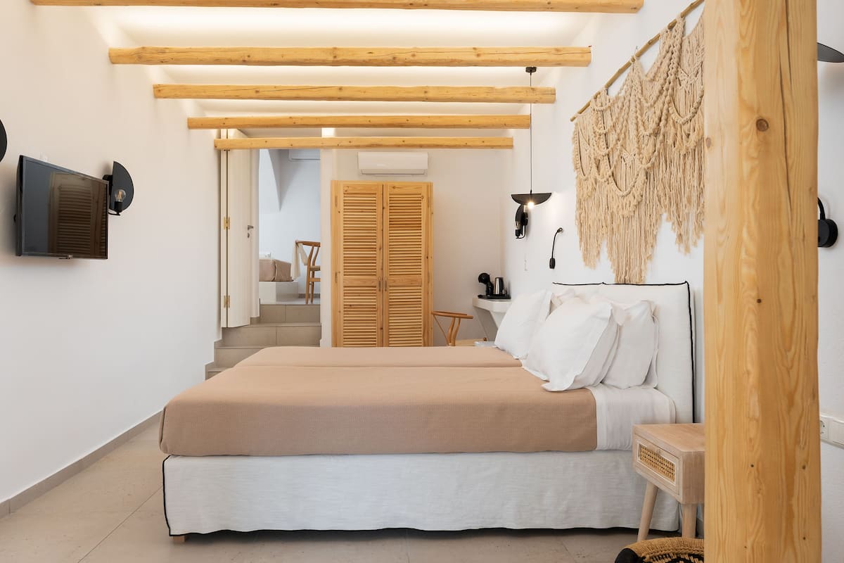 A bedroom with a bed, wooden beams, a television, a macrame wall hanging, and a staircase leading to another room.