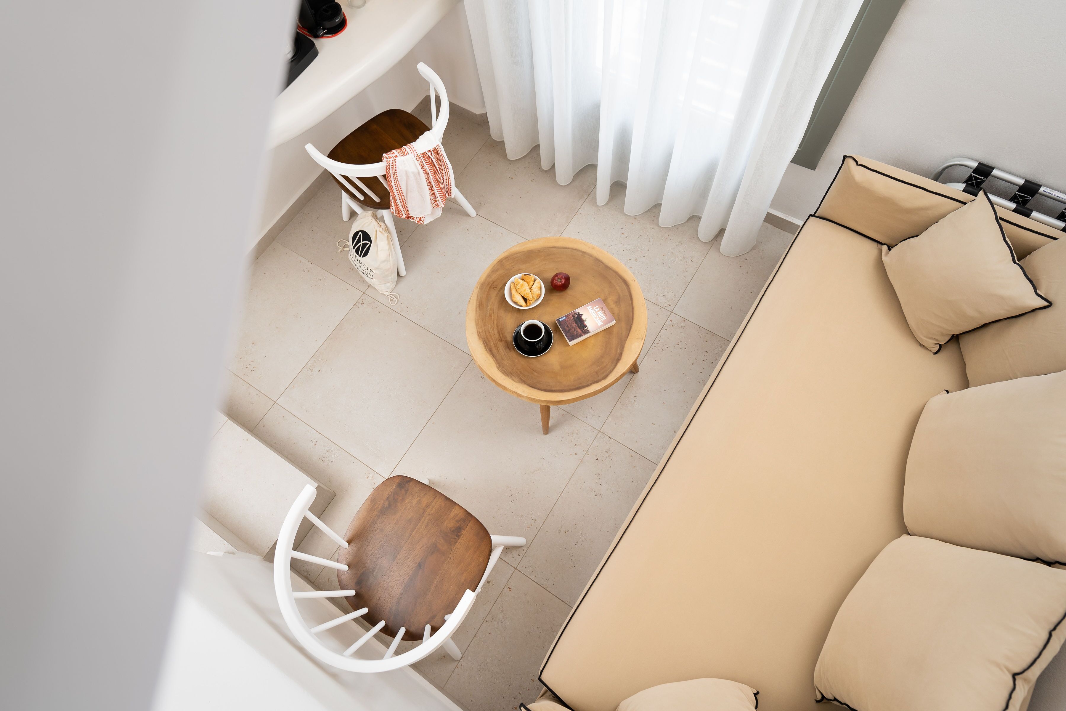 Uma sala de estar moderna com um sofá bege, uma mesa de centro de madeira, uma cadeira branca e uma mesinha com um livro e uma xícara.