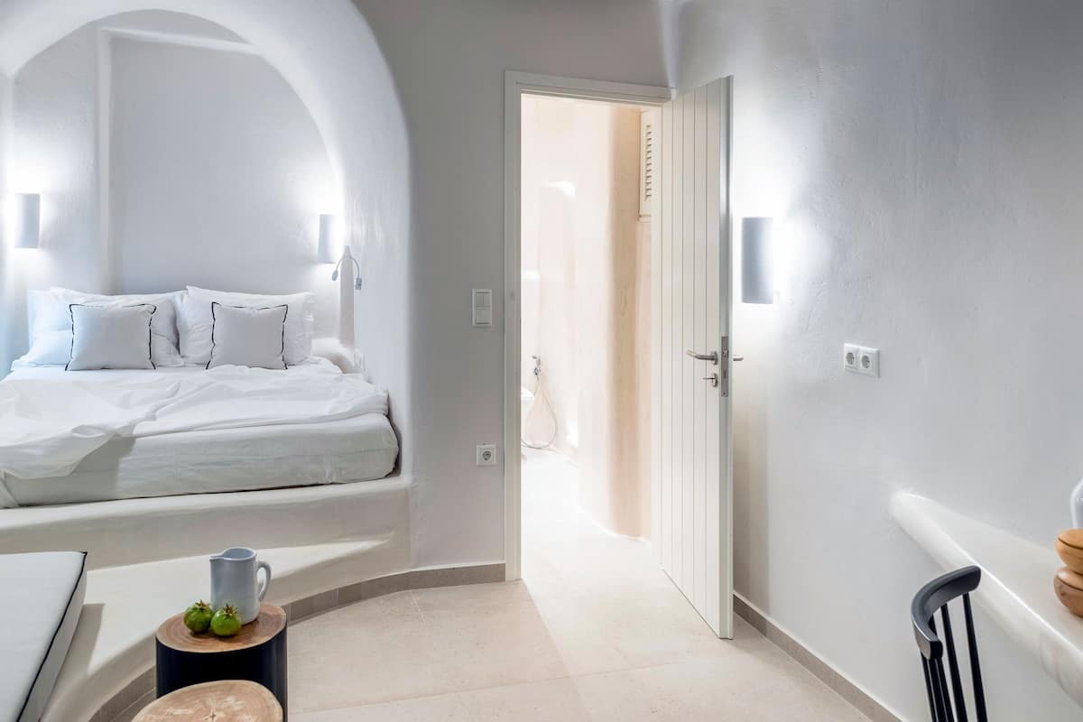 A bedroom with a bed, a bedside table, a wooden stool, and a door leading to another room.