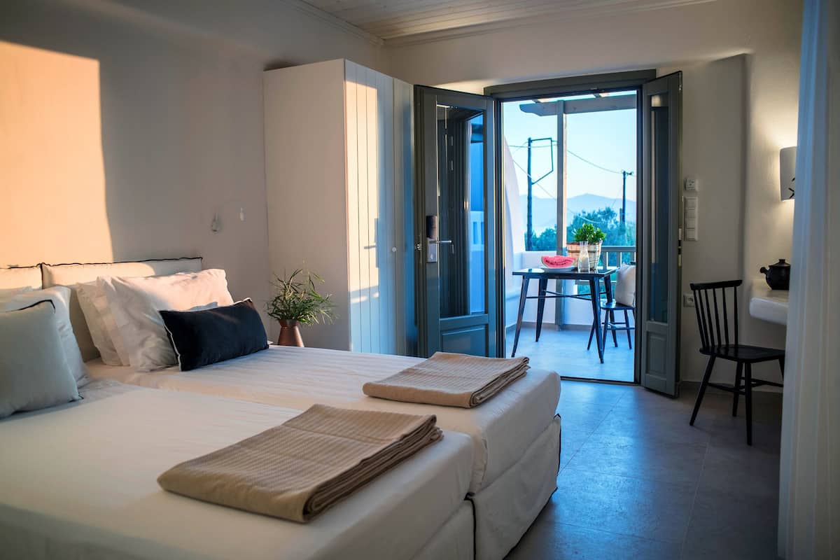 A bedroom with a bed, pillows, a bedside table, a potted plant, and a view of an outdoor dining area through an open door.