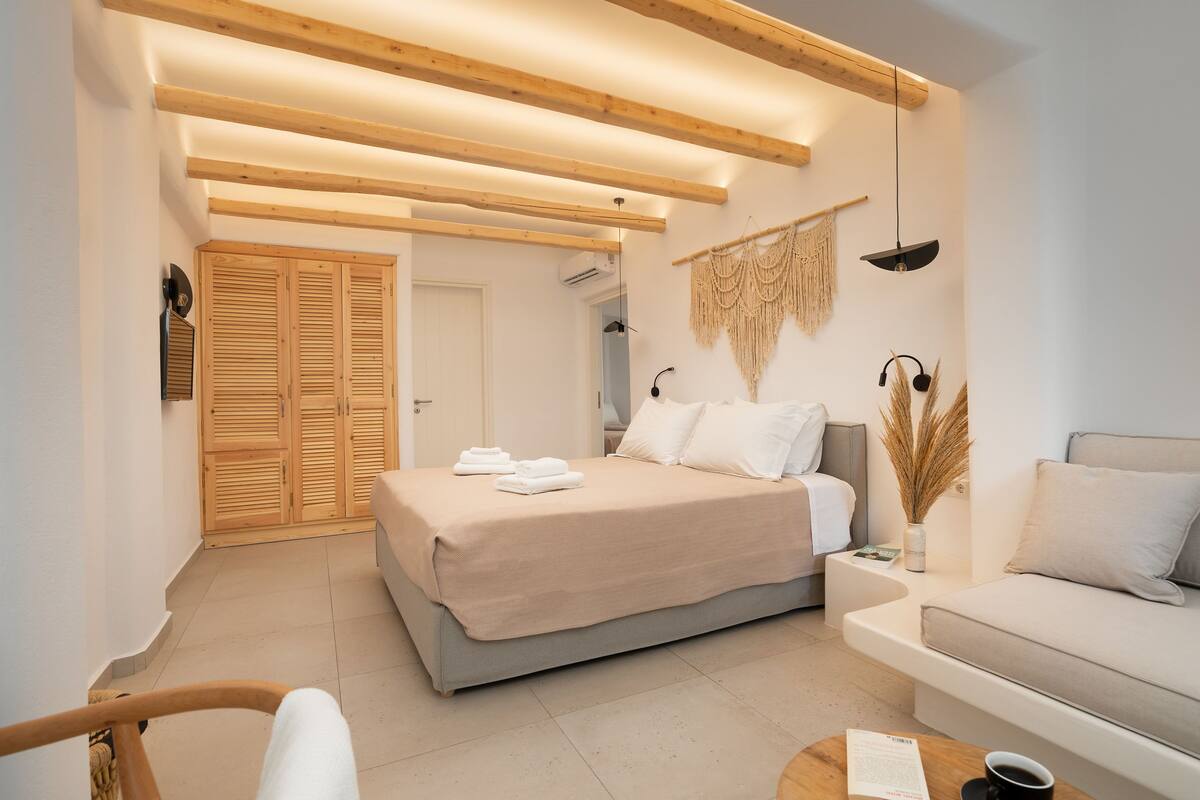 A modern bedroom with a bed, a sofa, and a coffee table. There is a wooden ceiling and a wall-mounted lamp.