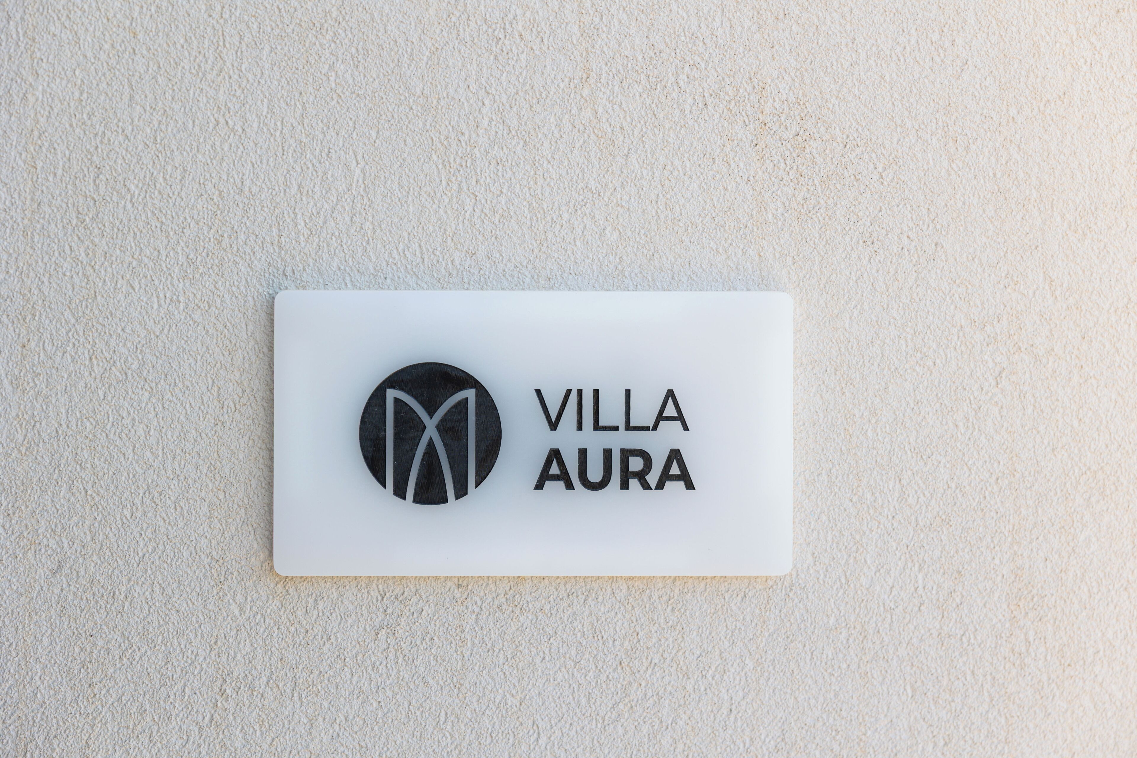 A white sign with the text 'VILLA AURA' and a logo, mounted on a textured wall.