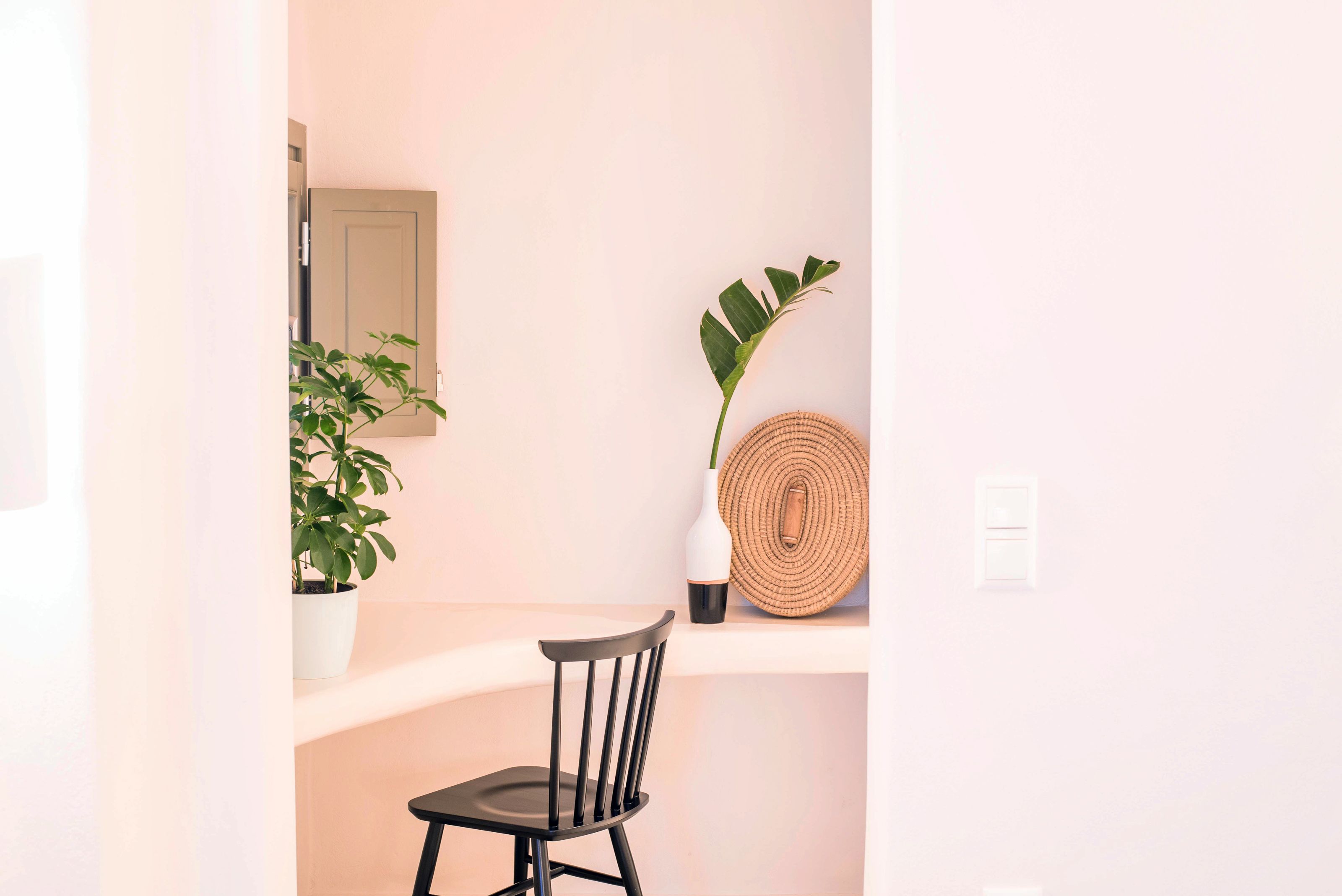 A minimalist room with a black chair, a potted plant, a woven wall hanging, and a small mirror.