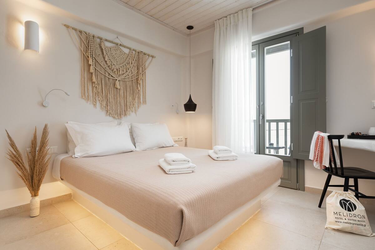 A neatly made bed with white linens, a macrame wall hanging, a bedside lamp, and a bag with the hotel name.