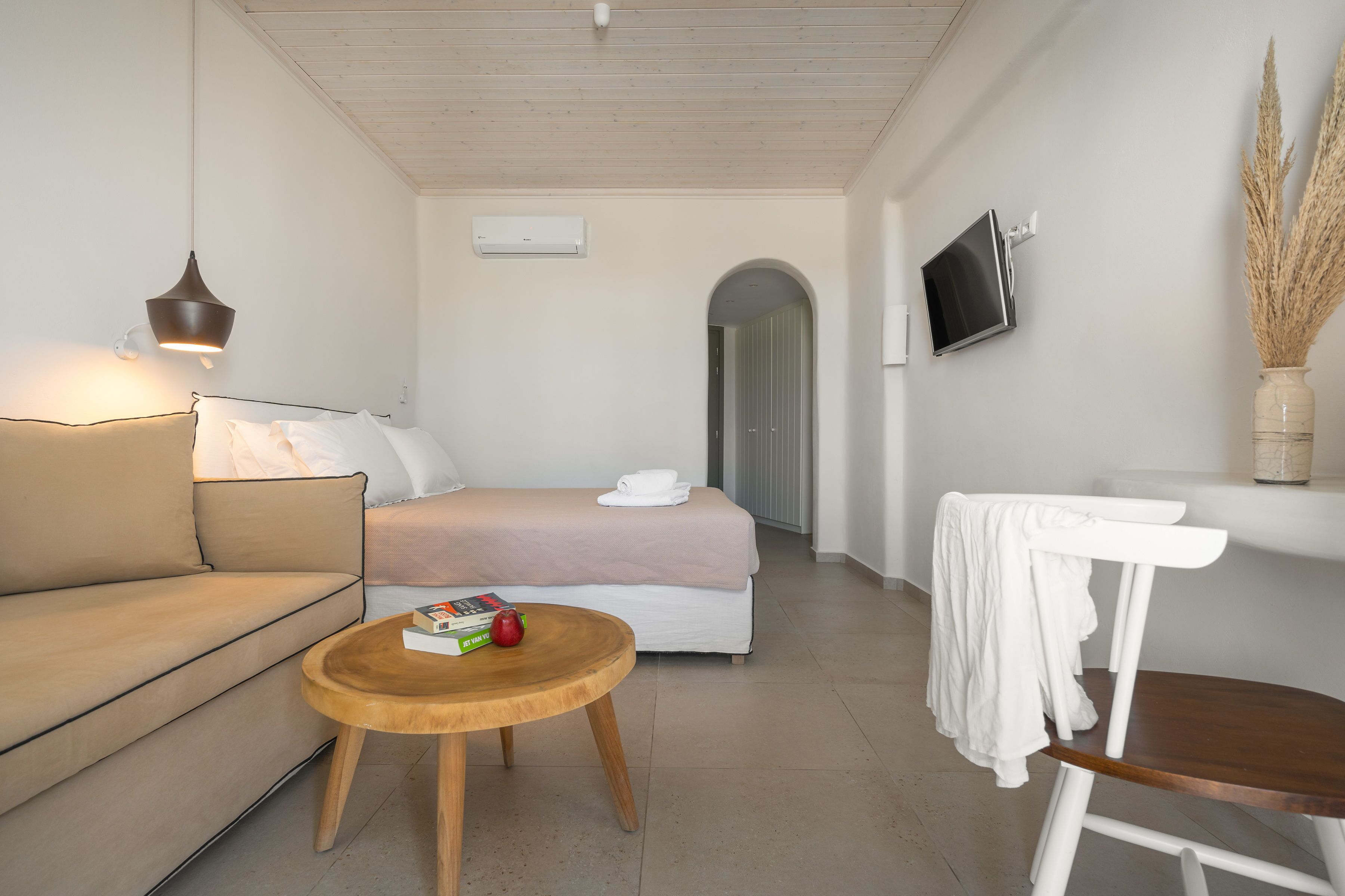 A modern hotel room with a bed, a sofa, a wooden table, a TV, and a vase with dried plants.