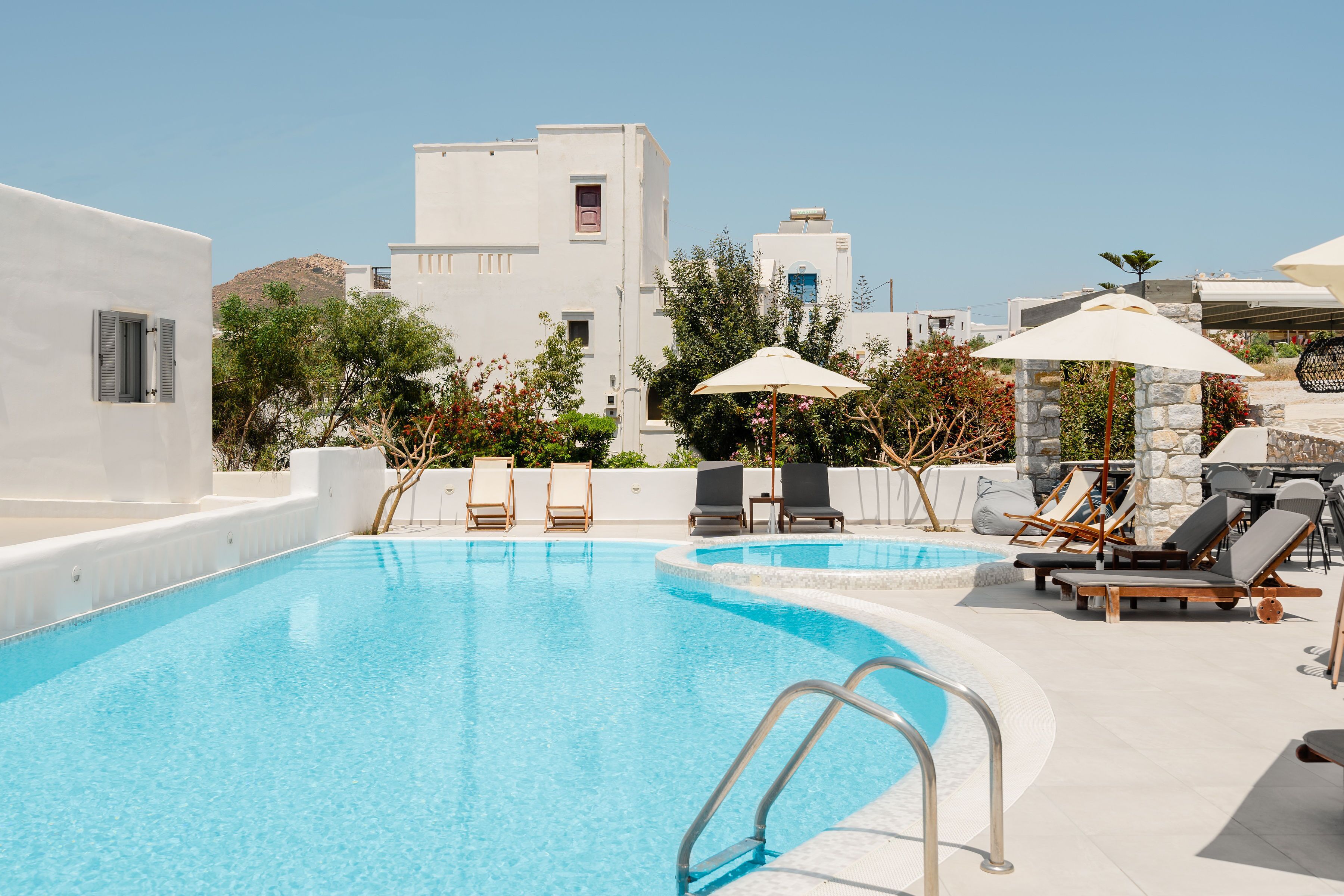 A swimming pool with lounge chairs and umbrellas, surrounded by white buildings and greenery.