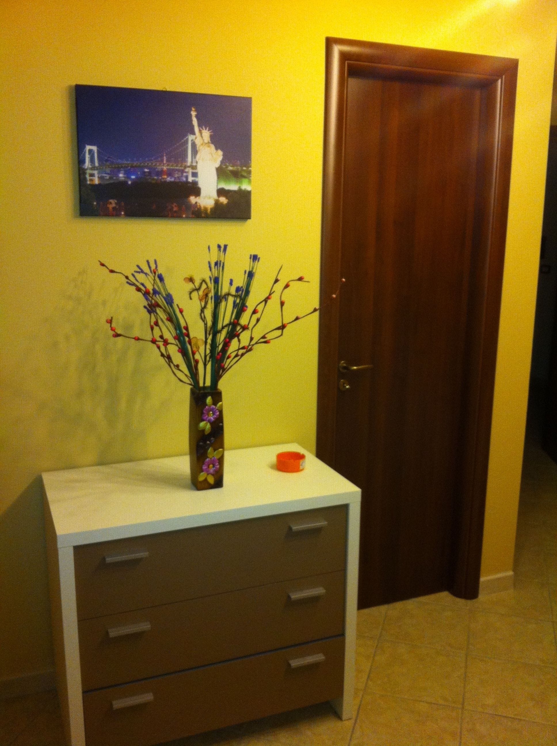 A room with a white dresser, a vase with branches, a framed picture of a bridge, and a closed wooden door.