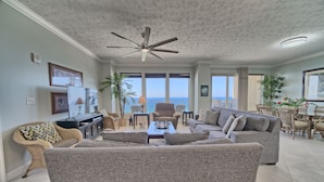 A spacious living room with a ceiling fan, a large gray sofa, a coffee table, and a dining area with wicker furniture. - Luxurious Beachfront Condo with Beachside Pool Access and Free WiFi - Unit 1202 by RedAwning (Panama City Beach)