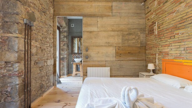 Een slaapkamer met een houten vloer, een bed met wit beddengoed, een houten hoofdbord en een bakstenen muur.