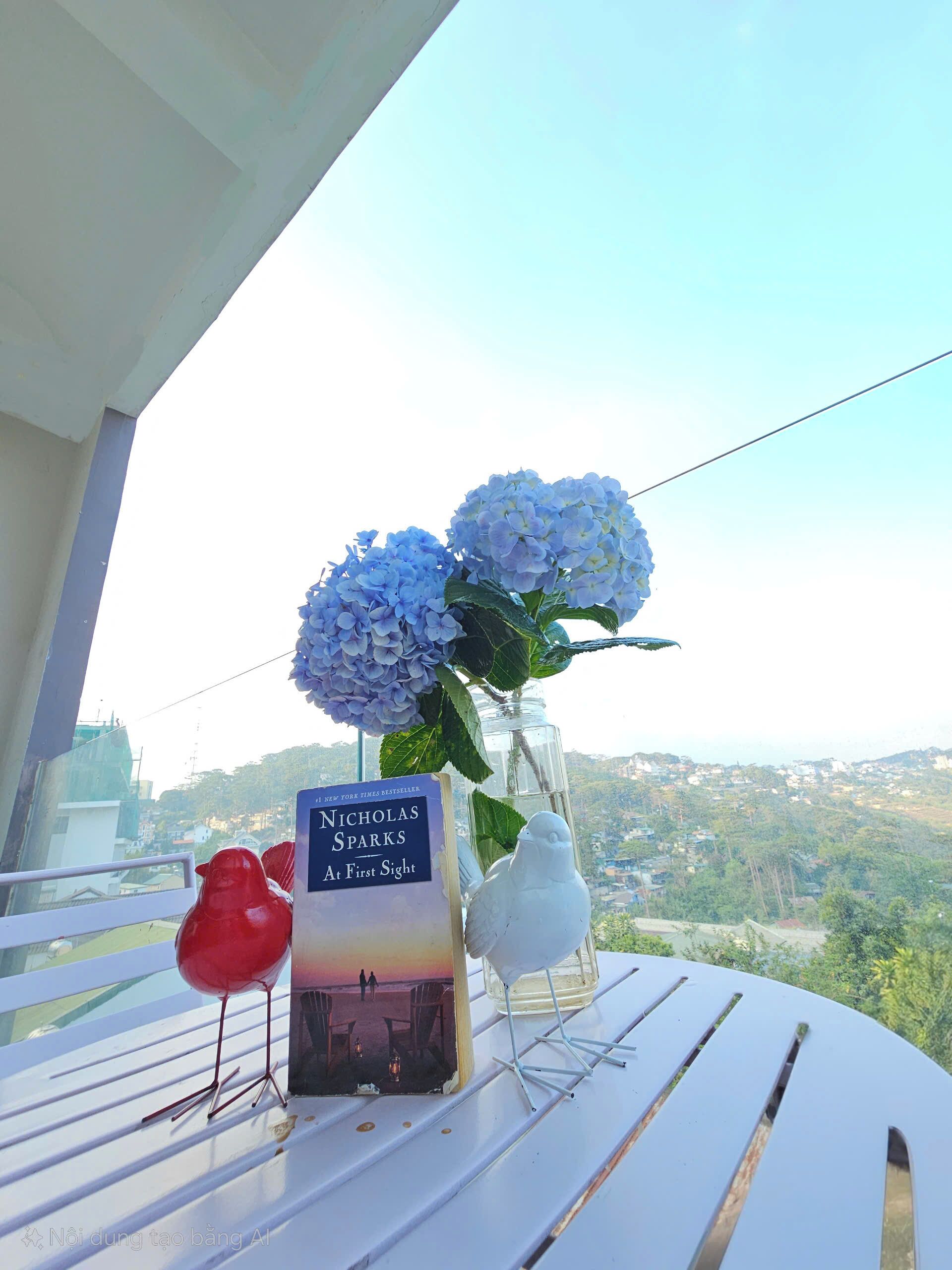 Une table sur laquelle se trouve un livre intitulé « Nicholas Sparks, le coup de foudre », un vase avec des fleurs bleues et une statuette d’oiseau blanche.