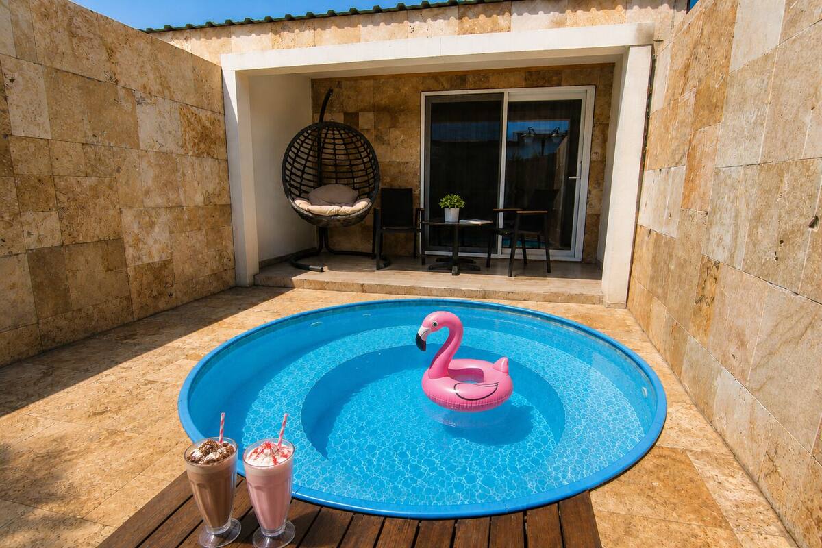 Een zwembad met een roze flamingo drijver, twee drankjes op het dek, en een stoel en tafel op de achtergrond.