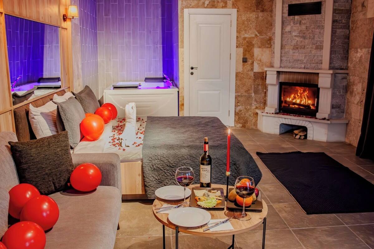 Een gezellige woonkamer met een open haard, een bank, een eettafel gedekt met een fles wijn, en rode ballonnen.
