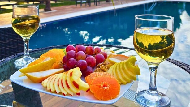 Mesa junto a la piscina con una bandeja de frutas y dos copas de vino blanco, rodeada de palmeras y una piscina.