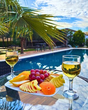 Mesa junto a la piscina con una bandeja de frutas y dos copas de vino blanco, rodeada de palmeras y una piscina.