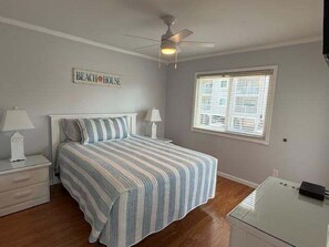 A bedroom with a bed, nightstands, a ceiling fan, a window with blinds, and a sign on the wall.