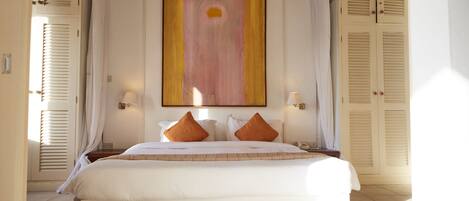 A large bed with white linens and two orange pillows, flanked by two bedside tables with lamps, in a room with a large abstract painting on the wall and two closed doors with shutters.