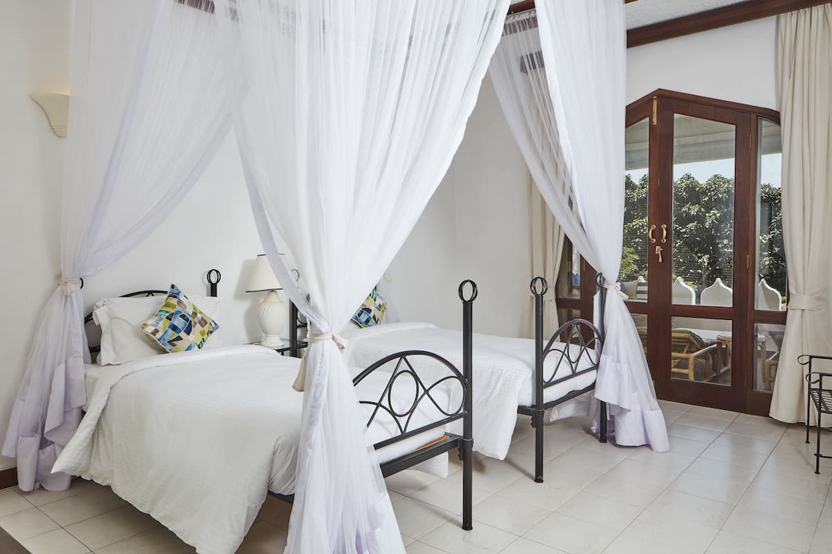 A bedroom with two beds, each with a canopy, a bedside table with a lamp, and a view of the outdoors through a glass door.