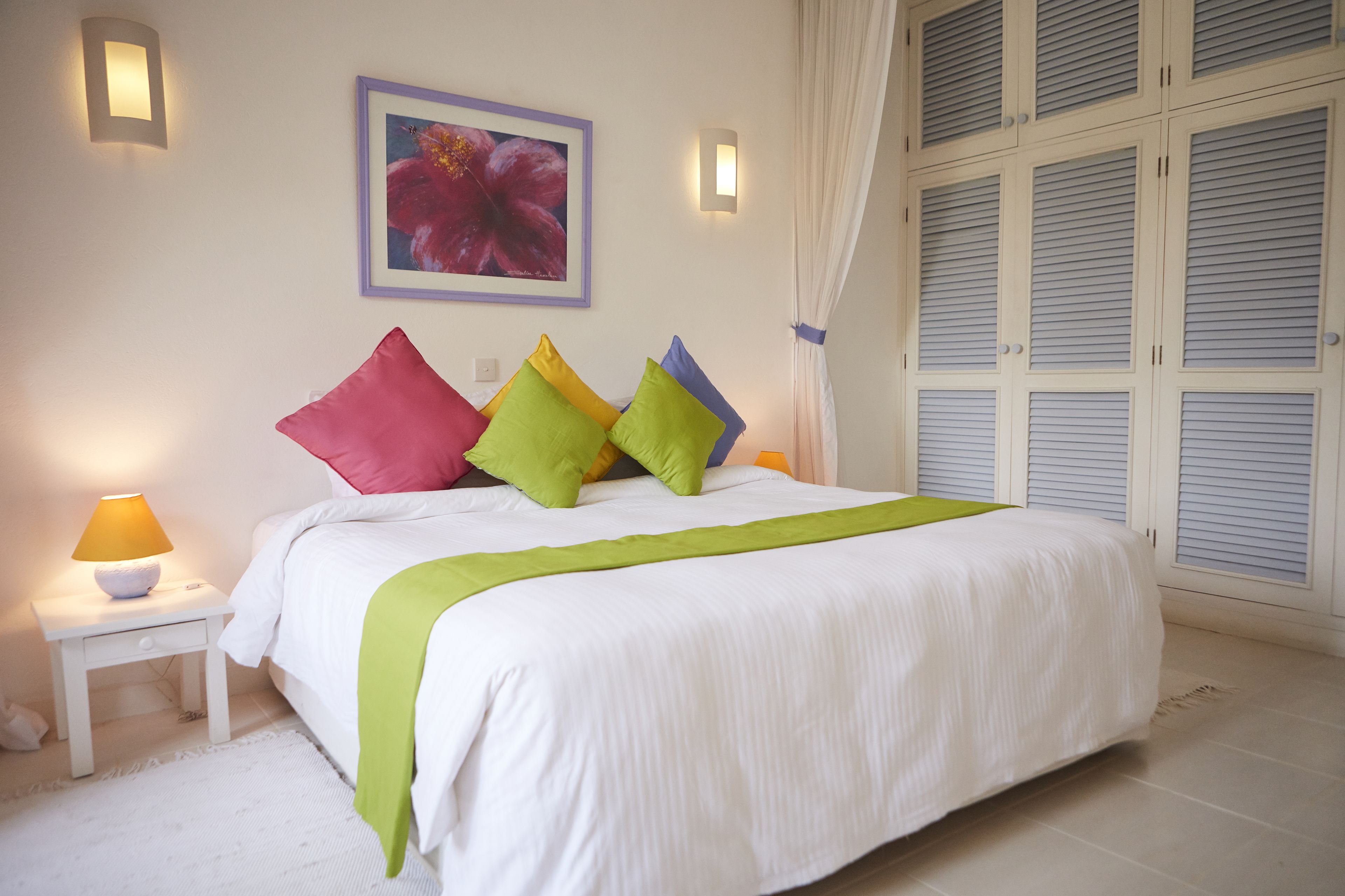 Cama bem arrumada com edredom branco, almofadas verdes e amarelas, e uma mesa de cabeceira branca com abajur.