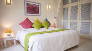 Cama bem arrumada com edredom branco, almofadas verdes e amarelas, e uma mesa de cabeceira branca com abajur.