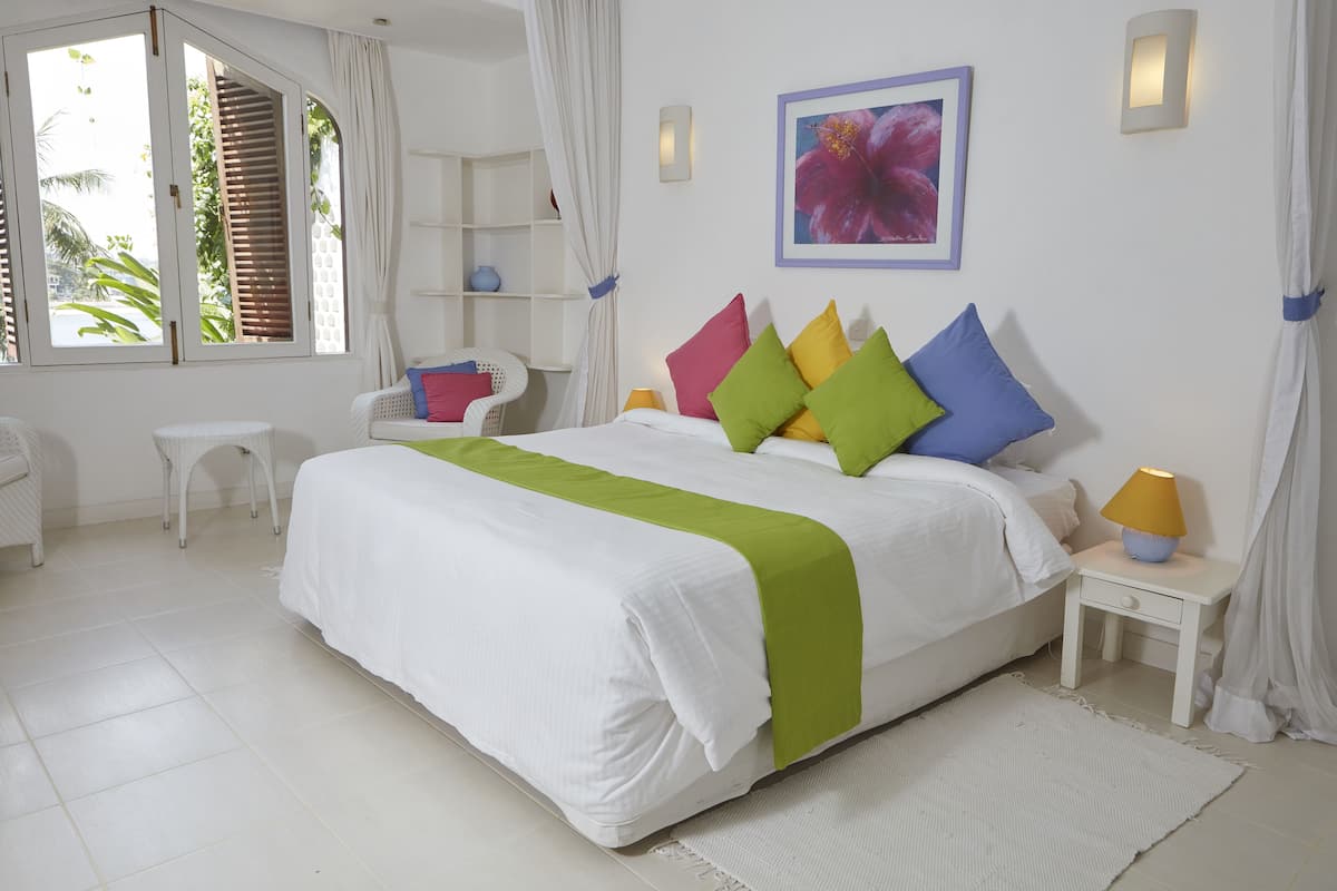 A neatly arranged bedroom with a large bed, a bedside table with a lamp, a small chair, and a window with curtains.