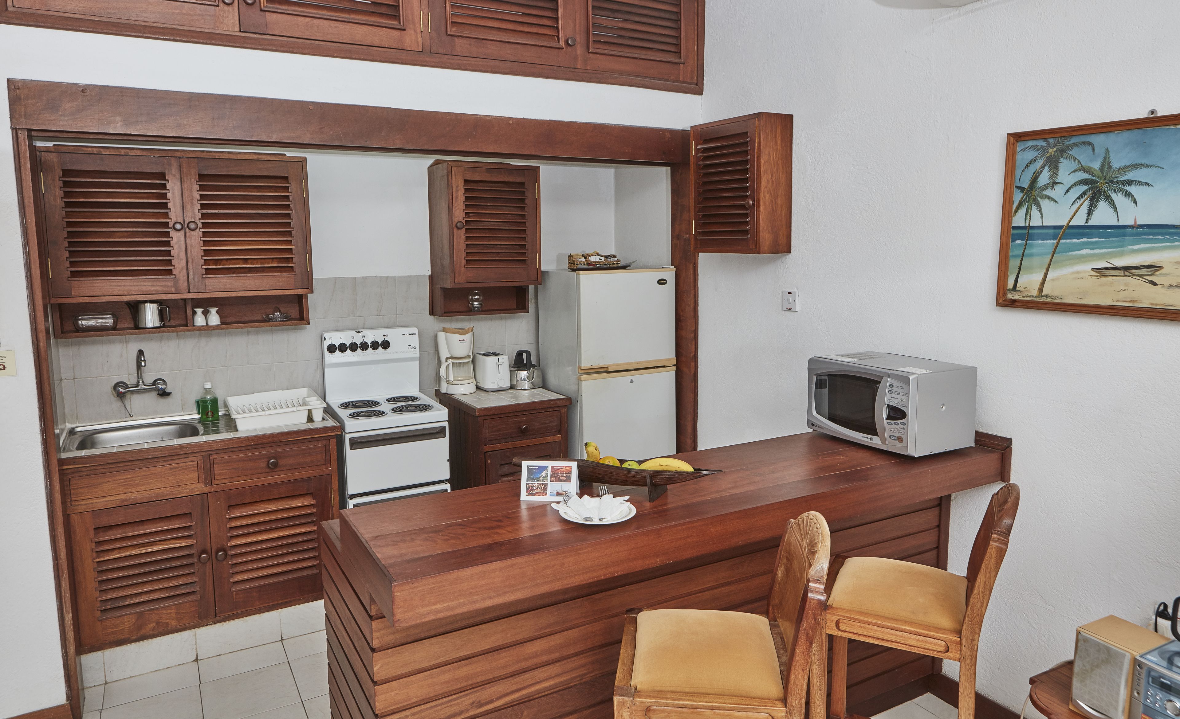 A compact kitchen with wooden cabinets, a stove, a refrigerator, and a microwave. There are two wooden chairs and a small table with a banana and a plate.