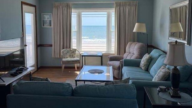 Habitación con sofá, sillón y una pequeña mesa. Se ve una ventana con cortinas, una lámpara y un televisor.