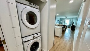 A laundry area with two stacked front-loading washers and dryers, a kitchen area with a dining table and chairs, and a hallway leading to a living space. - Boston Commons 102 3 Bedroom Condo by RedAwning (Surfside Beach)