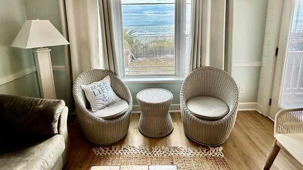 A room with wicker chairs, a small round table, and a view of the beach through a window. - Boston Commons 102 3 Bedroom Condo by RedAwning (Surfside Beach)