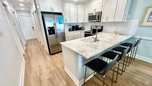 A modern kitchen with white cabinets, stainless steel appliances, a marble countertop, and bar stools. - Boston Commons 102 3 Bedroom Condo by RedAwning (Surfside Beach)