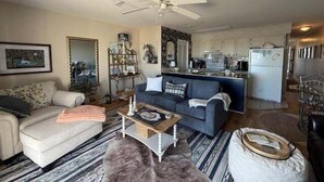 A living room with a sofa, coffee table, and a ceiling fan. There is a kitchen area in the background.