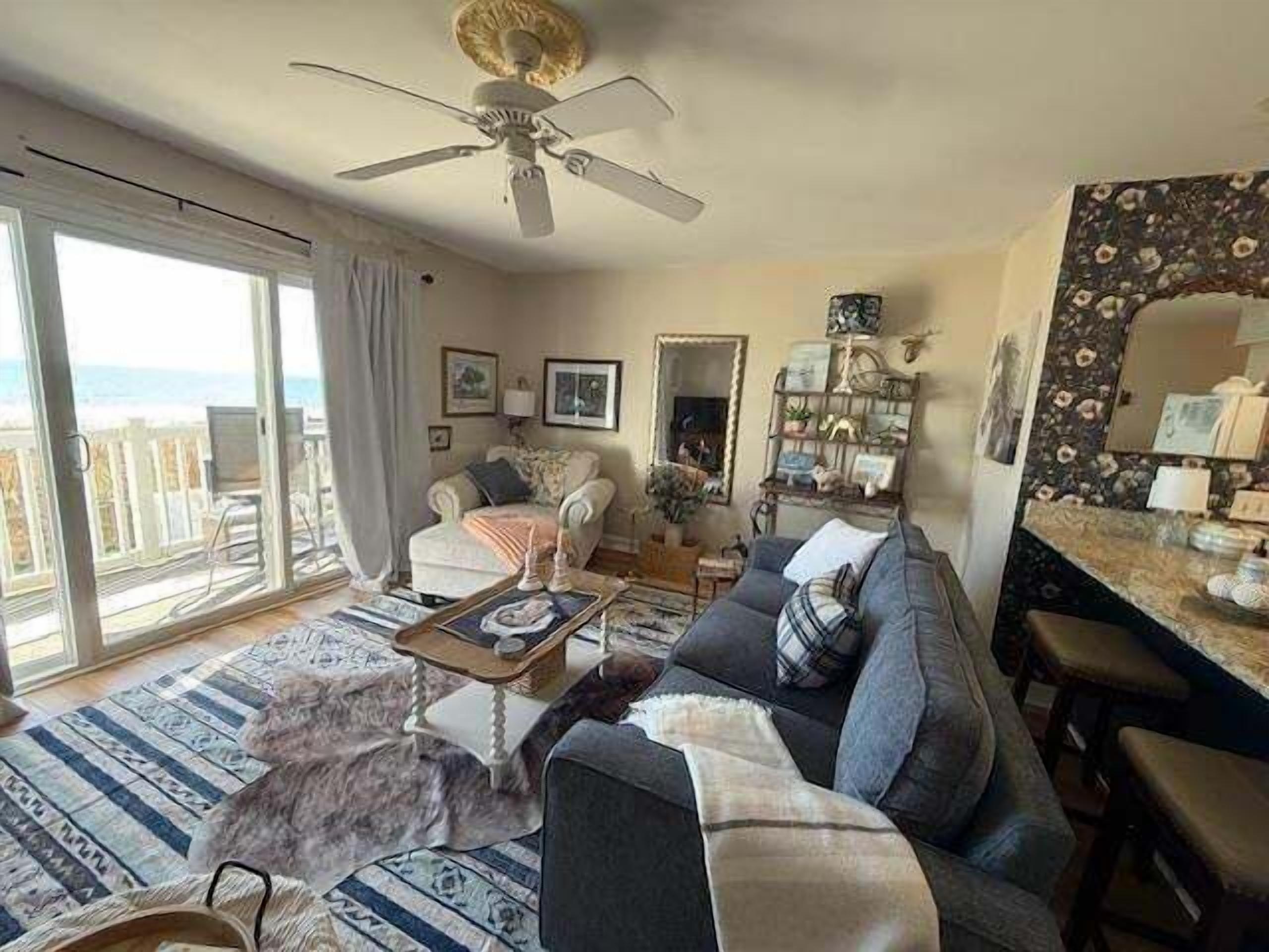 Een woonkamer met een bank, een salontafel en een hond. Er is een plafondventilator, een raam met uitzicht en een spiegel aan de muur.