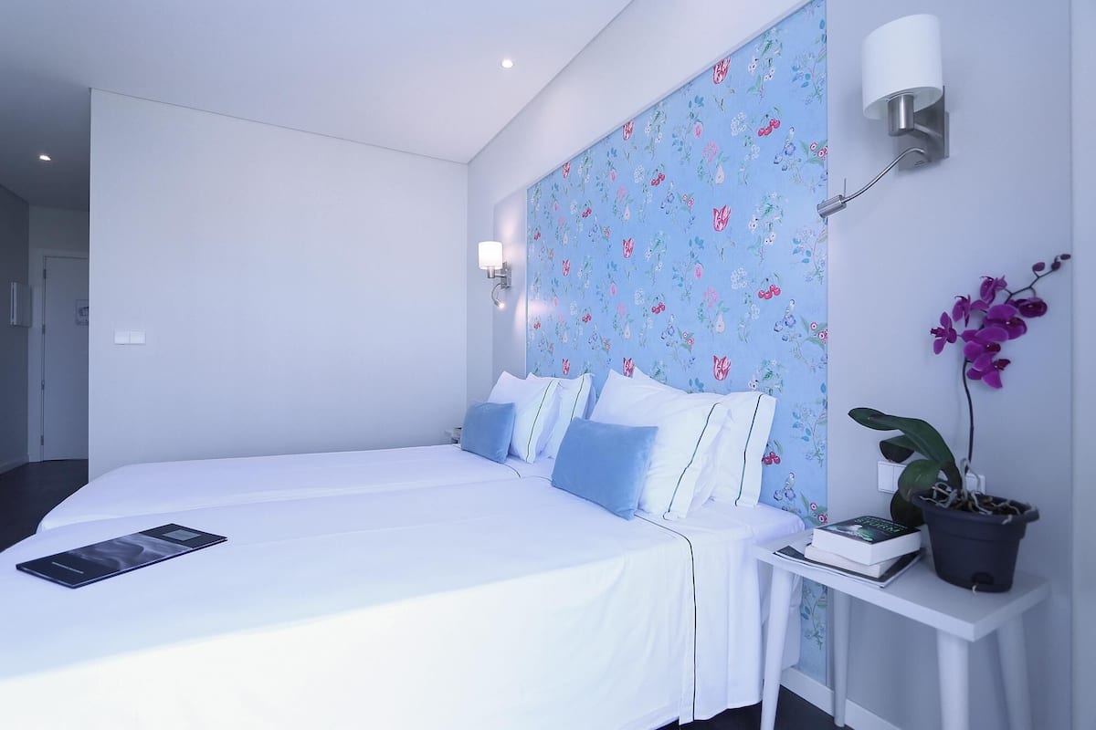 Une chambre à coucher avec un lit, une table de chevet, une orchidée en pot et un mur recouvert d’un papier peint à motifs floraux.