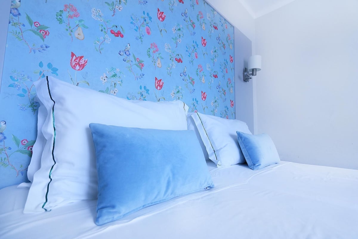 Un lit avec une literie blanche et des oreillers bleus, placé devant un mur orné d’un motif floral.
