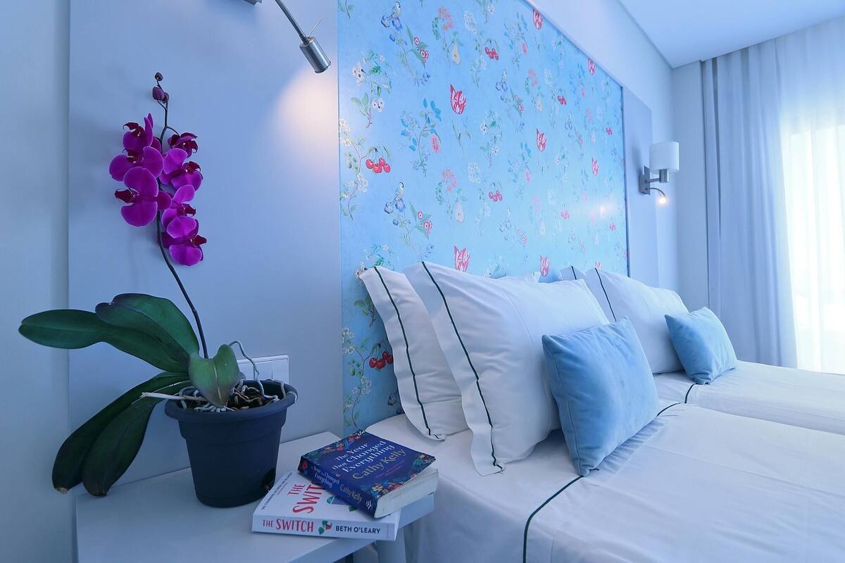 Une chambre d’hôtel moderne, équipée d’un lit, d’une table de chevet, d’une plante en pot et d’un mur recouvert d’un papier peint à motifs floraux.