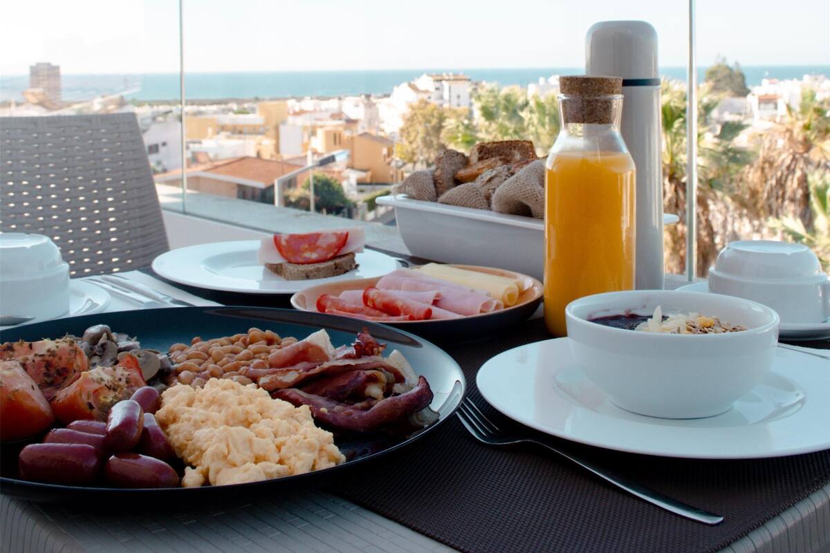 Une table de petit-déjeuner avec différents plats, une bouteille de jus d’orange et une vue sur la ville et la mer.
